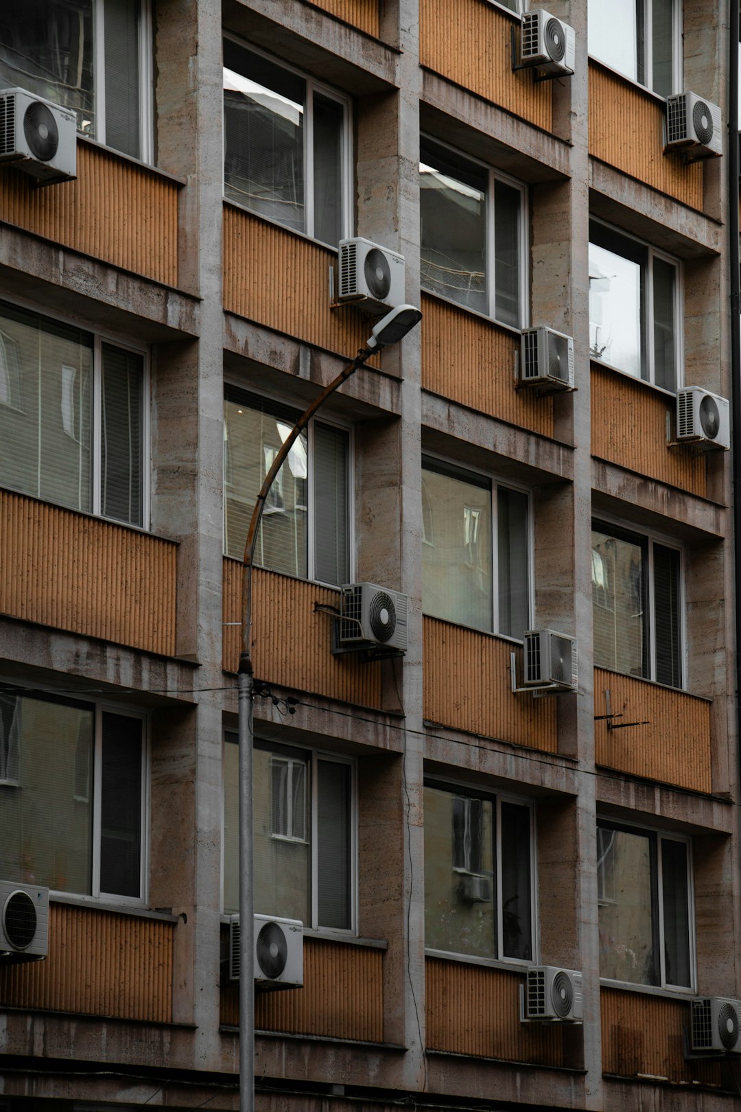 An urban apartment building with multiple air conditioning units mounted on the facade, showcasing a repetitive pattern of windows and industrial design elements. This image highlights the everyday architecture of city life, emphasizing the functional aspects of residential buildings.